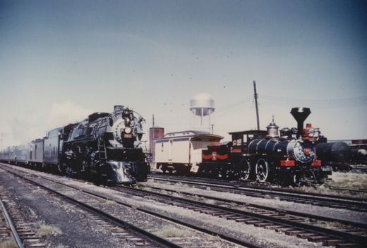 Two steam locomotives, one large and black with the number 6032 and one small and black with the number 35, are on parallel tracks in a railroad yard with a white passenger car and a water tower in the background.