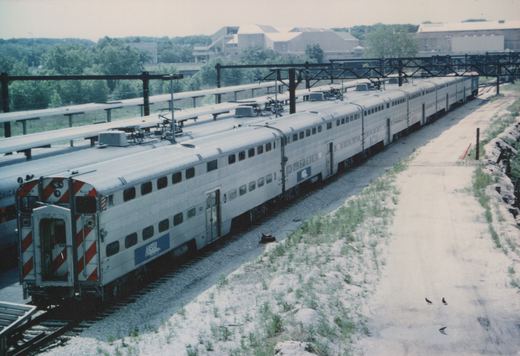 A silver Metra passenger train with blue lettering is parked on tracks under overhead wires, viewed from a high angle on a cloudy day.