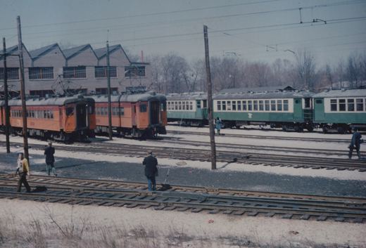 A winter scene in a rail yard showing two South Shore interurban trains, one orange and one green, on parallel tracks with several people on the ground nearby.