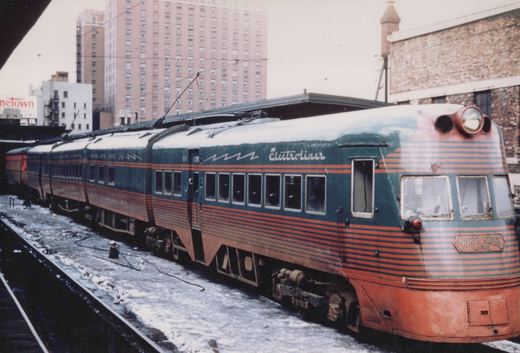A green and orange streamlined electric train, labeled Electroliner and North Shore Line, stands at an elevated station platform with snow on the ground.