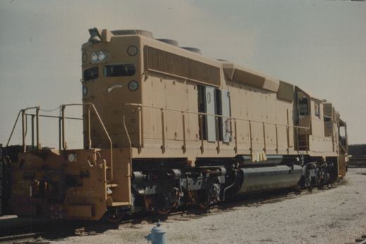 A side view of a tan diesel locomotive with a long hood, parked on tracks in a gravel yard under a clear sky.