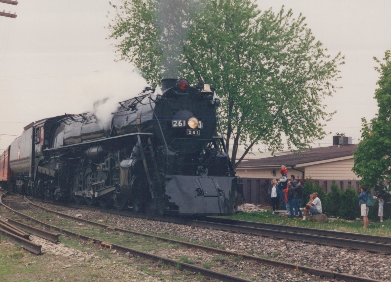 A black steam locomotive with the number 261 on its front and side is moving on tracks, with a group of people watching from the grass beside the tracks.