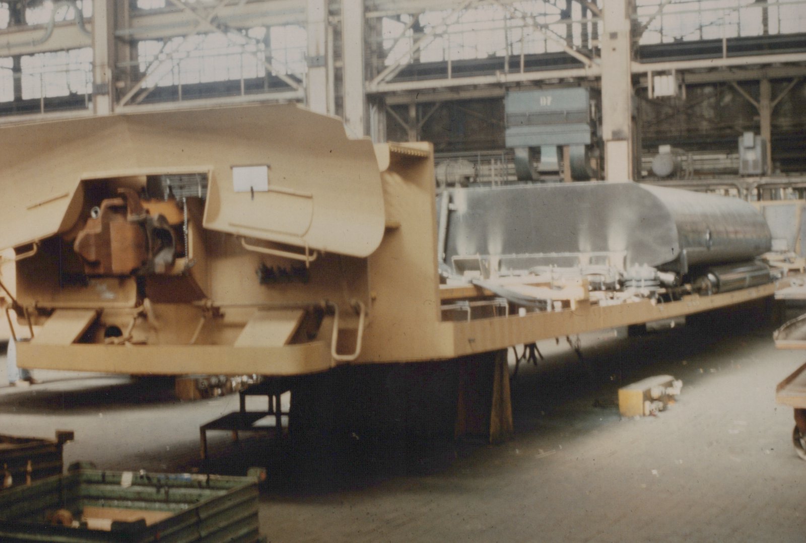 A large, tan-colored, partially assembled piece of rail equipment on a flatbed is being built inside a factory with high windows and steel beams, with another piece of machinery labeled 'D7' visible in the background.