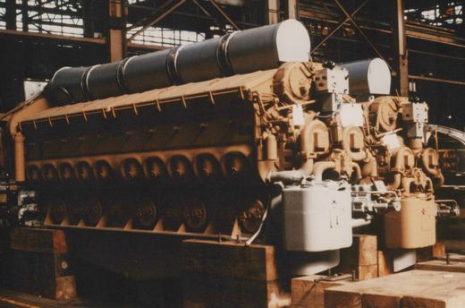 A large, unpainted diesel locomotive under construction inside a factory, resting on wooden blocks with its engine block and exhaust stacks visible.