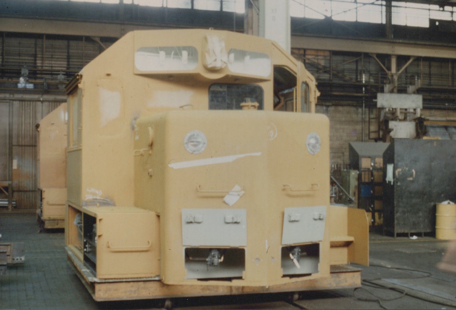 A yellow, unpainted diesel locomotive with a high short hood and covered windows sits inside a large industrial building, with workshop equipment in the background.