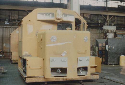A yellow, unpainted diesel locomotive with a high short hood and covered windows sits inside a large industrial building, with workshop equipment in the background.