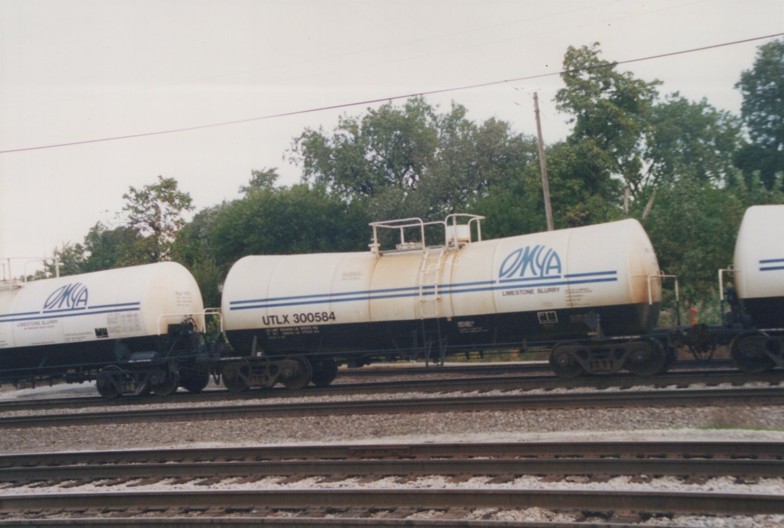 A side view of a white and black UTLX tank car with the number 300584 and OMYA branding, sitting on railroad tracks under an overcast sky with trees in the background.