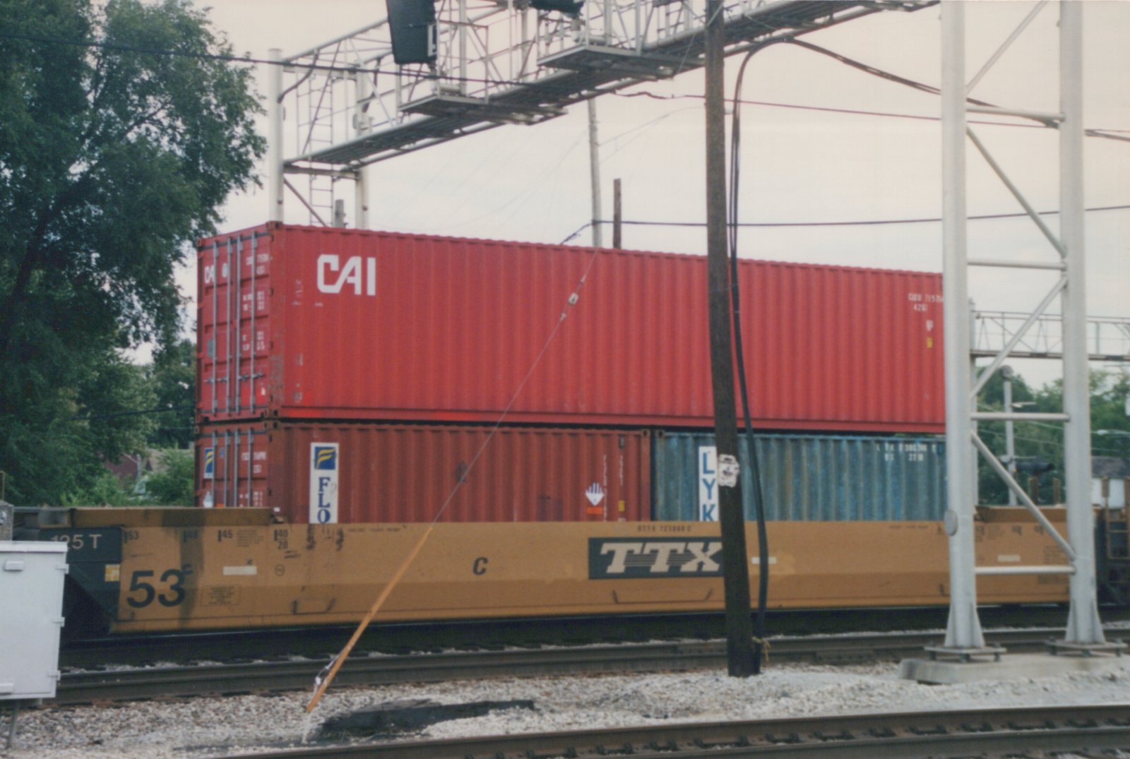 Side view of a yellow TTX freight car with the number 53, carrying three stacked shipping containers, under an overpass with overhead wires.
