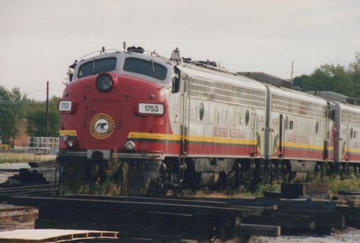 A three-quarter frontal shot of an Algoma Central diesel locomotive, number 1753, in a red and silver livery, sitting on tracks in a rail yard under an overcast sky.