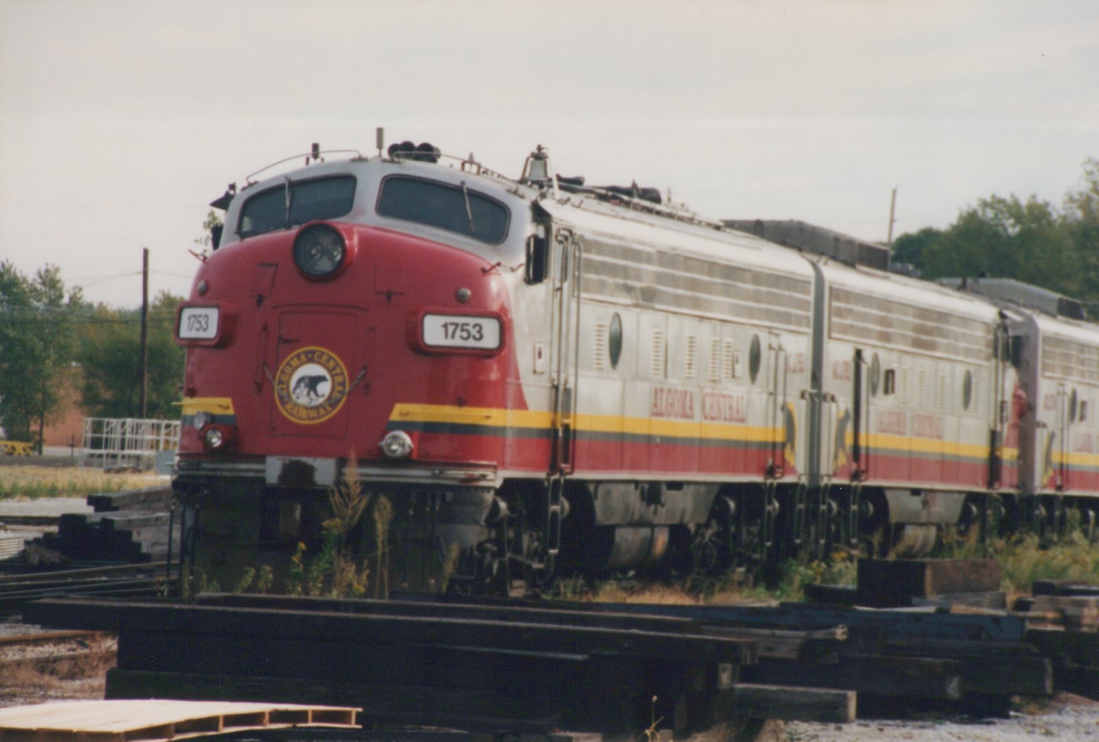 A three-quarter frontal shot of an Algoma Central diesel locomotive, number 1753, in a red and silver livery, sitting on tracks in a rail yard under an overcast sky.