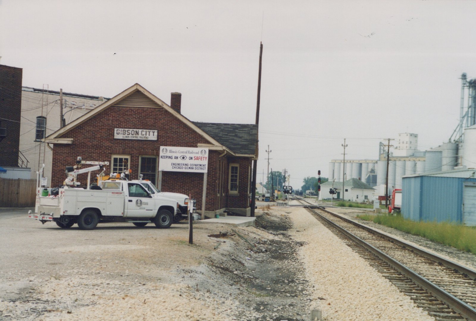A white Illinois Central Railroad utility truck is parked in front of the Gibson City station. A set of railroad tracks extends into the distance toward large grain silos under an overcast sky.