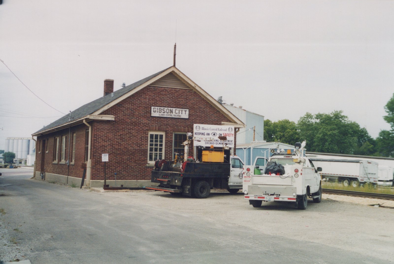 A brick railroad station with a sign reading 'Gibson City, Illinois Central Railroad' and two utility trucks parked in front under an overcast sky.