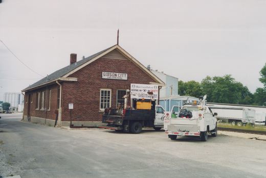A brick railroad station with a sign reading 'Gibson City, Illinois Central Railroad' and two utility trucks parked in front under an overcast sky.