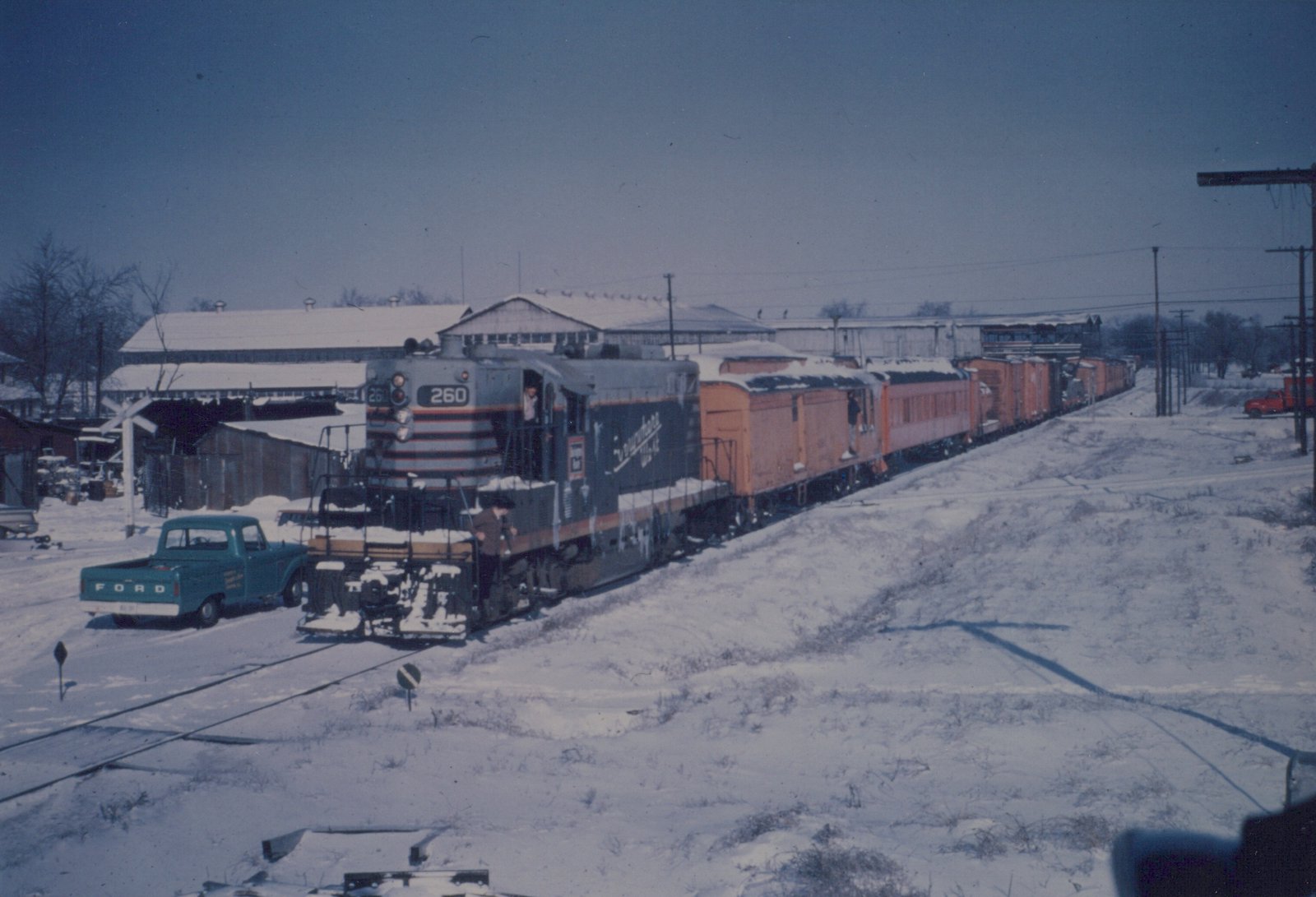 A diesel locomotive numbered 260, painted in Chicago Burlington and Quincy livery, leads a freight train through a snow-covered scene next to a blue Ford pickup truck.