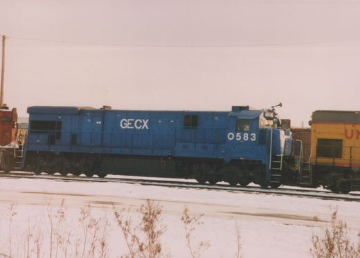A side view of a blue GECX diesel locomotive, number 0583, in a snow-covered rail yard with other locomotives partially visible.