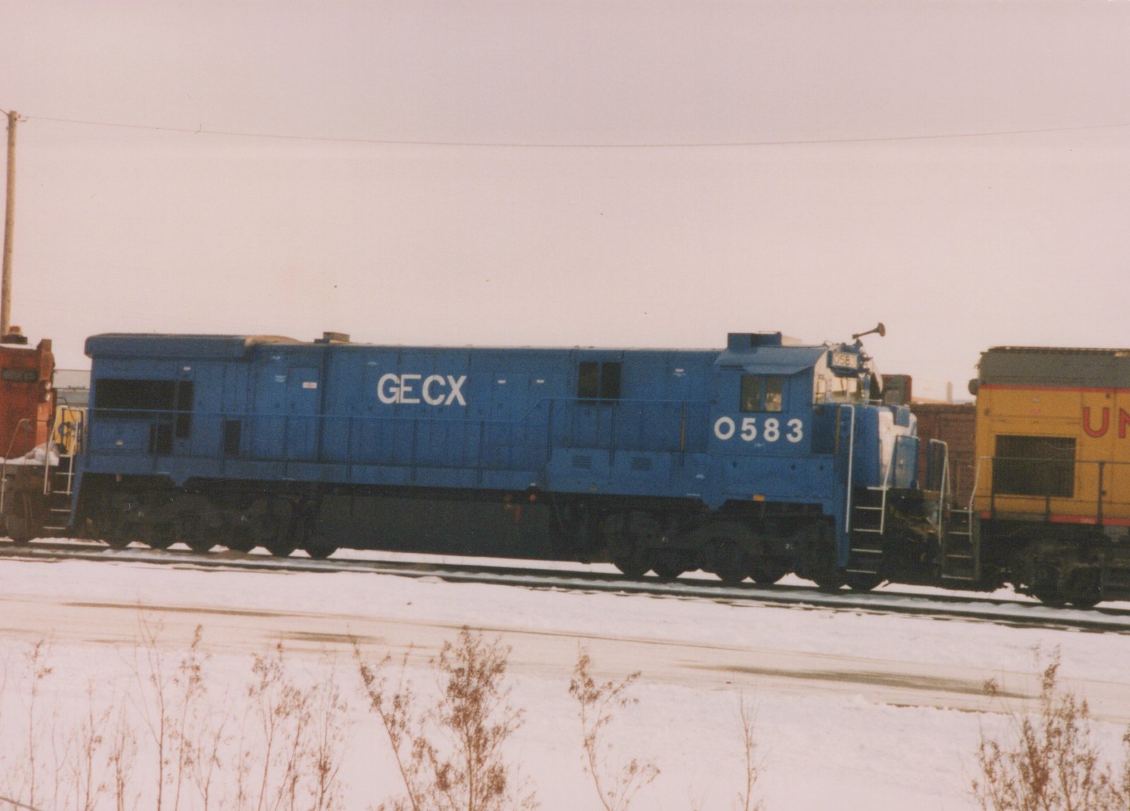 A side view of a blue GECX diesel locomotive, number 0583, in a snow-covered rail yard with other locomotives partially visible.