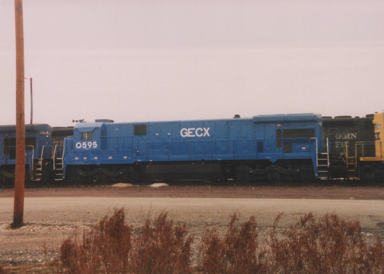 A side view of a blue GECX diesel locomotive numbered 0595, parked in a yard with other locomotives and dry brush in the foreground.