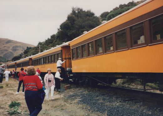 A side view of a group of people boarding a bright orange passenger train with brown trim, set against a backdrop of grassy hills and trees on an overcast day.