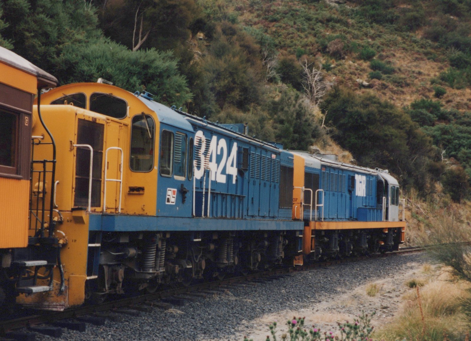 A side view of two blue and yellow diesel locomotives, numbered 3424 and 3407, with 'TGR' lettering, traveling on a railroad track next to a grassy, wooded hillside.