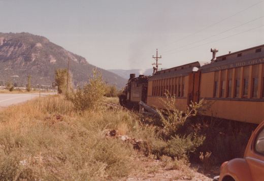 A steam locomotive with the number 576 and passenger car from the Denver & Rio Grande Western Railroad travel along tracks beside a highway, with a mountain in the background and a Volkswagen Beetle in the foreground.