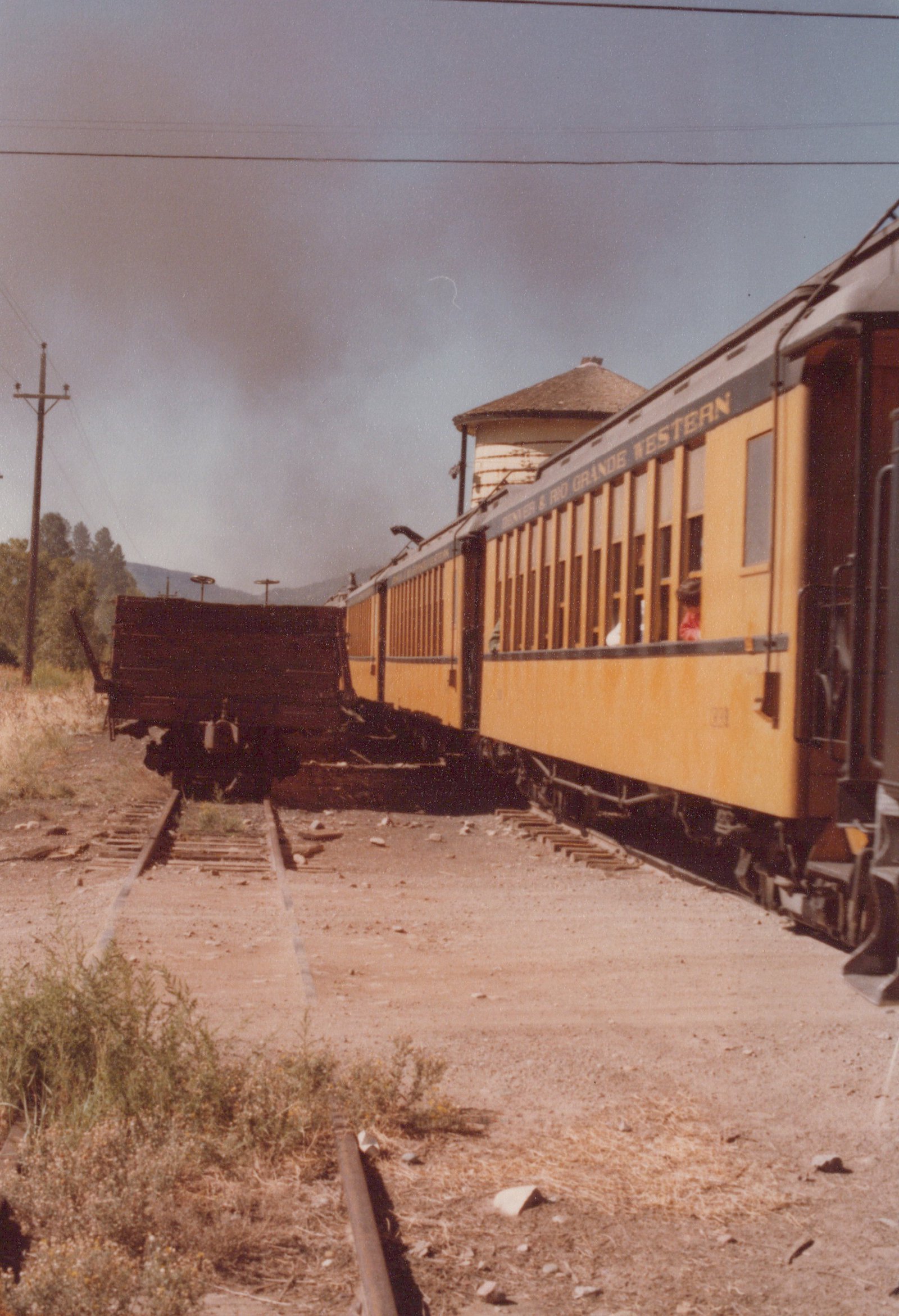A yellow Denver & Rio Grande Western passenger car is shown from a three-quarter rear view on a railroad track, with a water tower visible behind it and a dark freight car on an adjacent track.