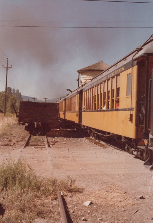 A yellow Denver & Rio Grande Western passenger car is shown from a three-quarter rear view on a railroad track, with a water tower visible behind it and a dark freight car on an adjacent track.