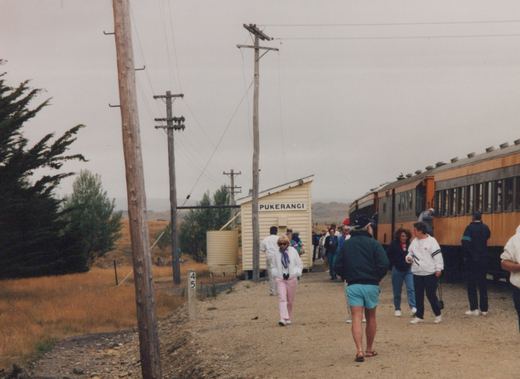 A group of people walking on a gravel platform beside a cream and brown train car at Pukerangi station, with wooden utility poles and a dry, grassy landscape in the background.