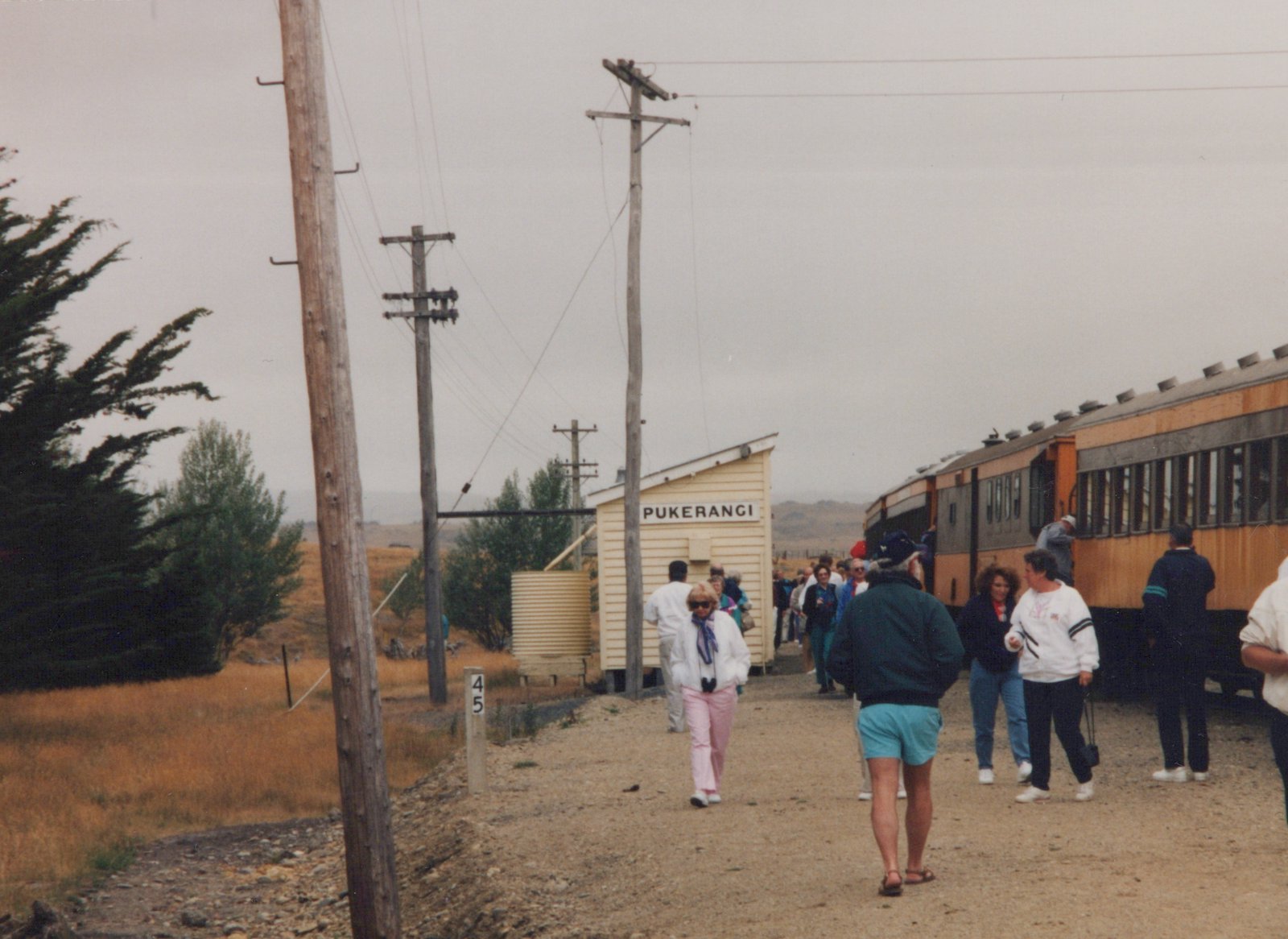 A group of people walking on a gravel platform beside a cream and brown train car at Pukerangi station, with wooden utility poles and a dry, grassy landscape in the background.