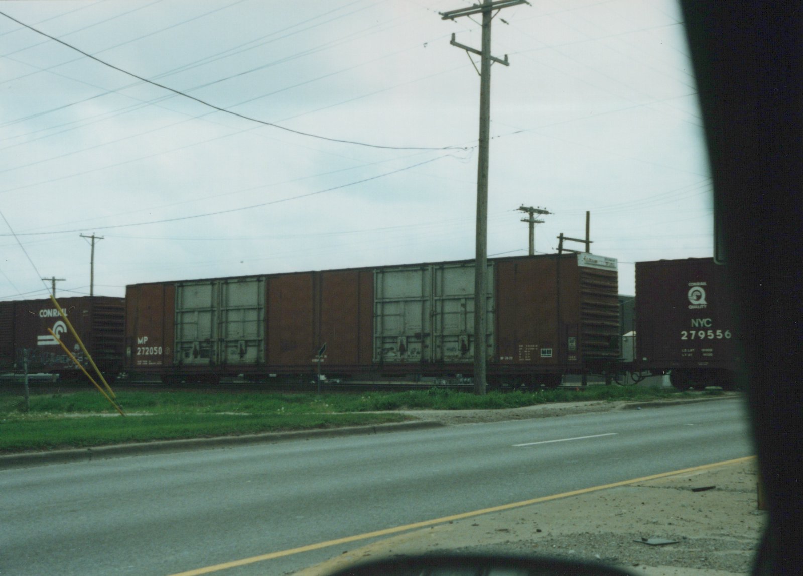 A photograph of a freight train with dark-colored boxcars on a track beside a road, viewed on an overcast day.