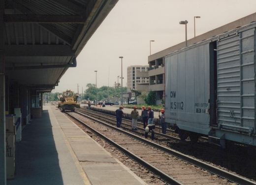 A gray freight car marked CNW X2 51112 is on the tracks beside a station platform, with several workers in hard hats on the tracks and a yellow maintenance vehicle nearby.