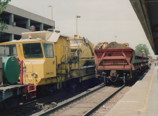 A yellow railroad work car labeled BUC 1 and CNWX 2001 is coupled to a red gondola with CNW 743002 and a flatcar with L 53 6, stationary at a station platform on a cloudy day.
