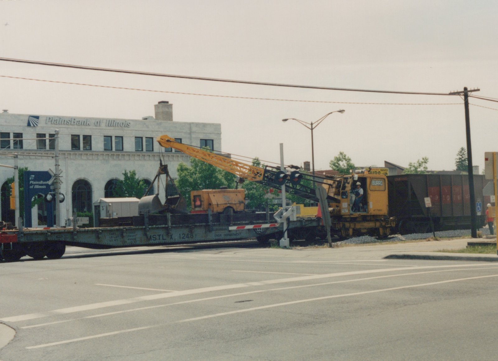 A yellow railroad crane mounted on a flatcar sits at a street-level railroad crossing, with a bank building visible in the background under an overcast sky.