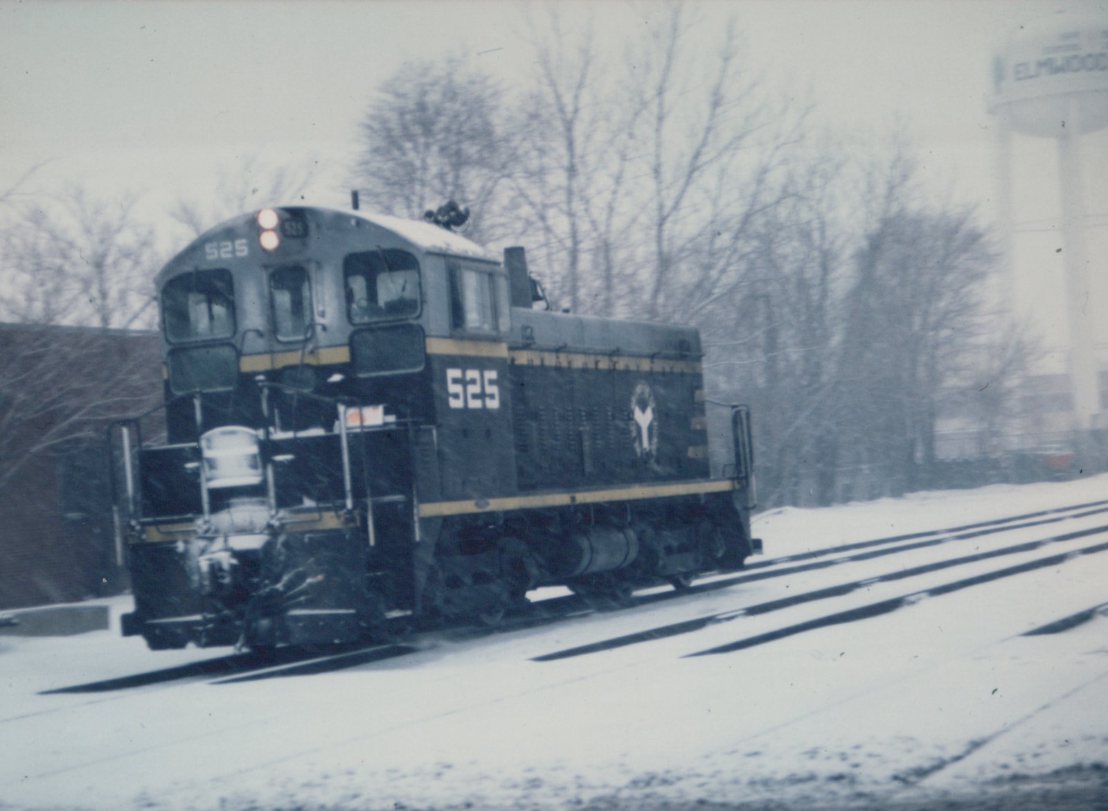A snow-covered EMD SW1200 diesel locomotive, number 525 with a white 'Y' logo, moves along tracks near a water tower marked 'ELMWOOD'.