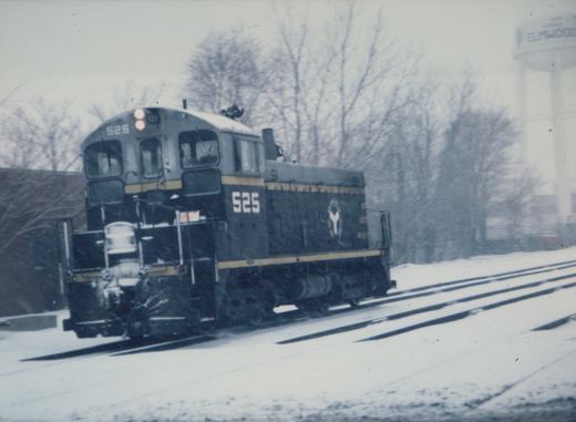 A snow-covered EMD SW1200 diesel locomotive, number 525 with a white 'Y' logo, moves along tracks near a water tower marked 'ELMWOOD'.