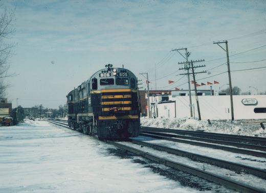 A gray and yellow C&NW diesel locomotive numbered 605 travels on snow-covered tracks, with utility poles and a white building in the background.