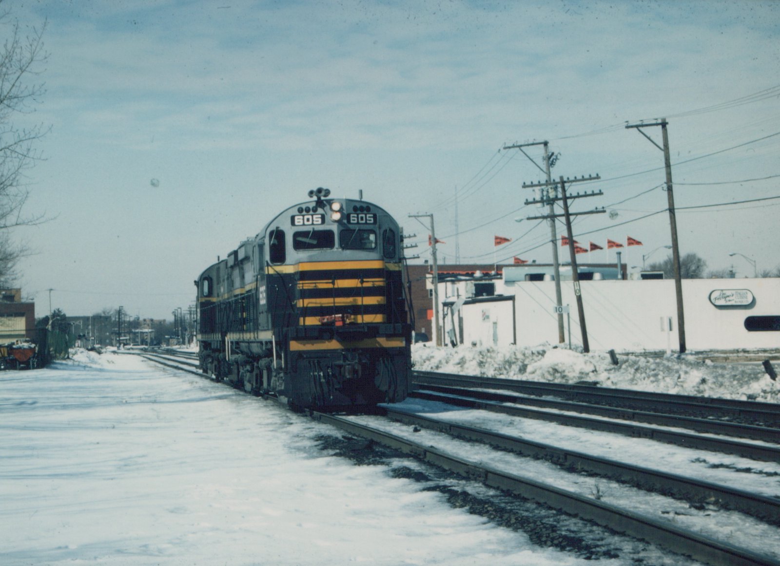 A gray and yellow C&NW diesel locomotive numbered 605 travels on snow-covered tracks, with utility poles and a white building in the background.