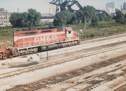 Side view of a red and white GM&O diesel locomotive numbered 908 on a track in a railyard, with a railroad bridge and city skyline in the background.
