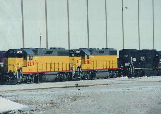 Three diesel locomotives are parked side by side in a snowy railyard: two yellow LLPX GP38-2s and one black Chicago and North Western GP40.