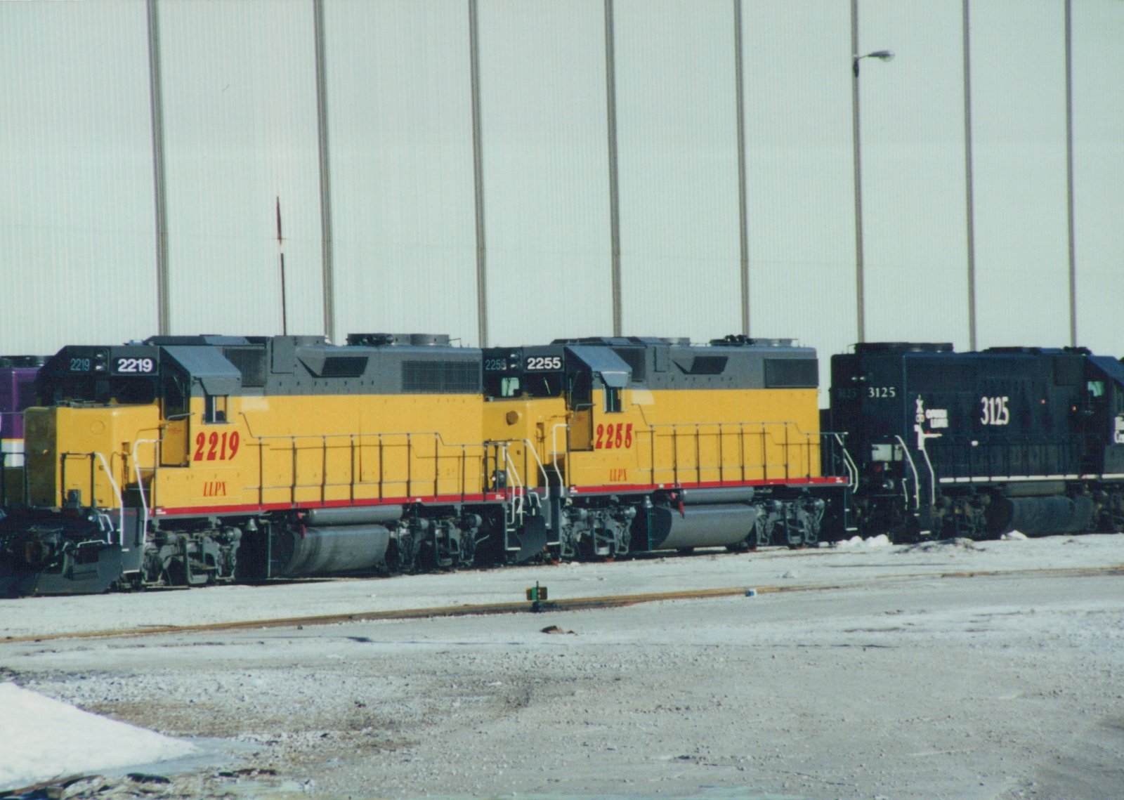 Three diesel locomotives are parked side by side in a snowy railyard: two yellow LLPX GP38-2s and one black Chicago and North Western GP40.