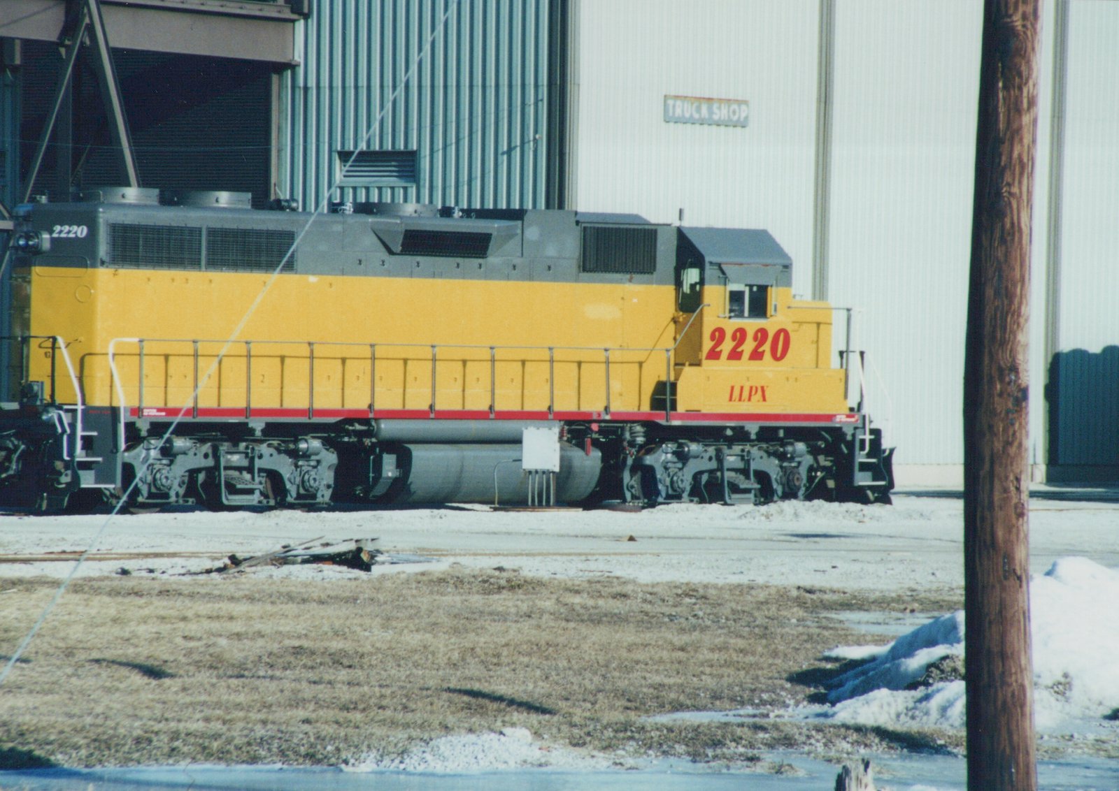 A yellow and gray diesel locomotive numbered 2220 with LLPX lettering stands on tracks in a snowy yard next to a building labeled 'TRUCK SHOP'.