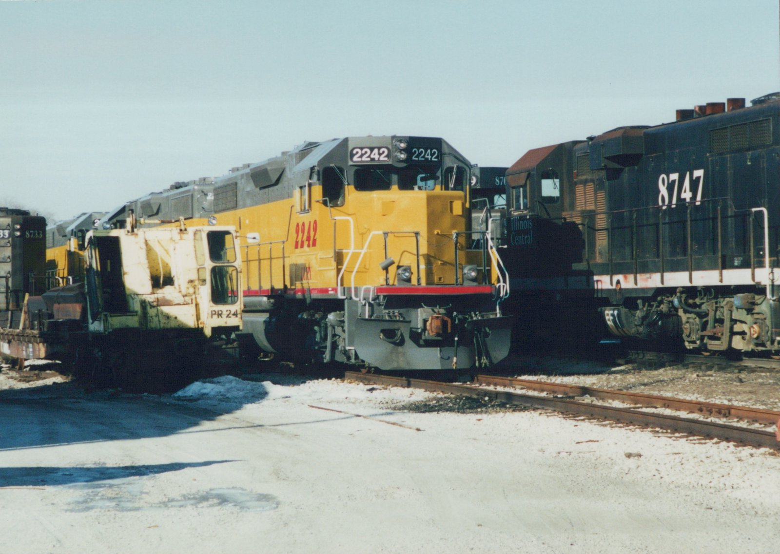 A yellow and gray Illinois Central GP38-2 locomotive numbered 2242 is parked on a track in a snowy yard, with black GP9 locomotives numbered 8747 and 8733 behind it.