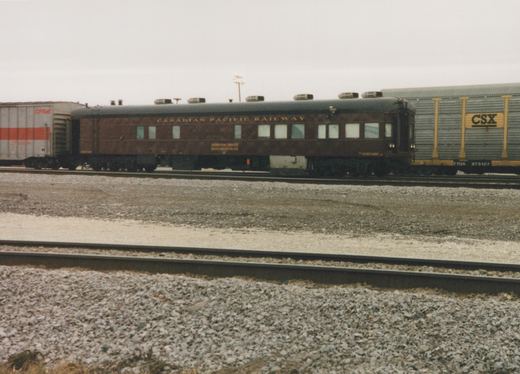 A side view of a brown Canadian Pacific passenger car labeled 'Track Evaluation Car 63' in a gravel rail yard, flanked by a grey CPRail boxcar and a grey CSX freight car under an overcast sky.
