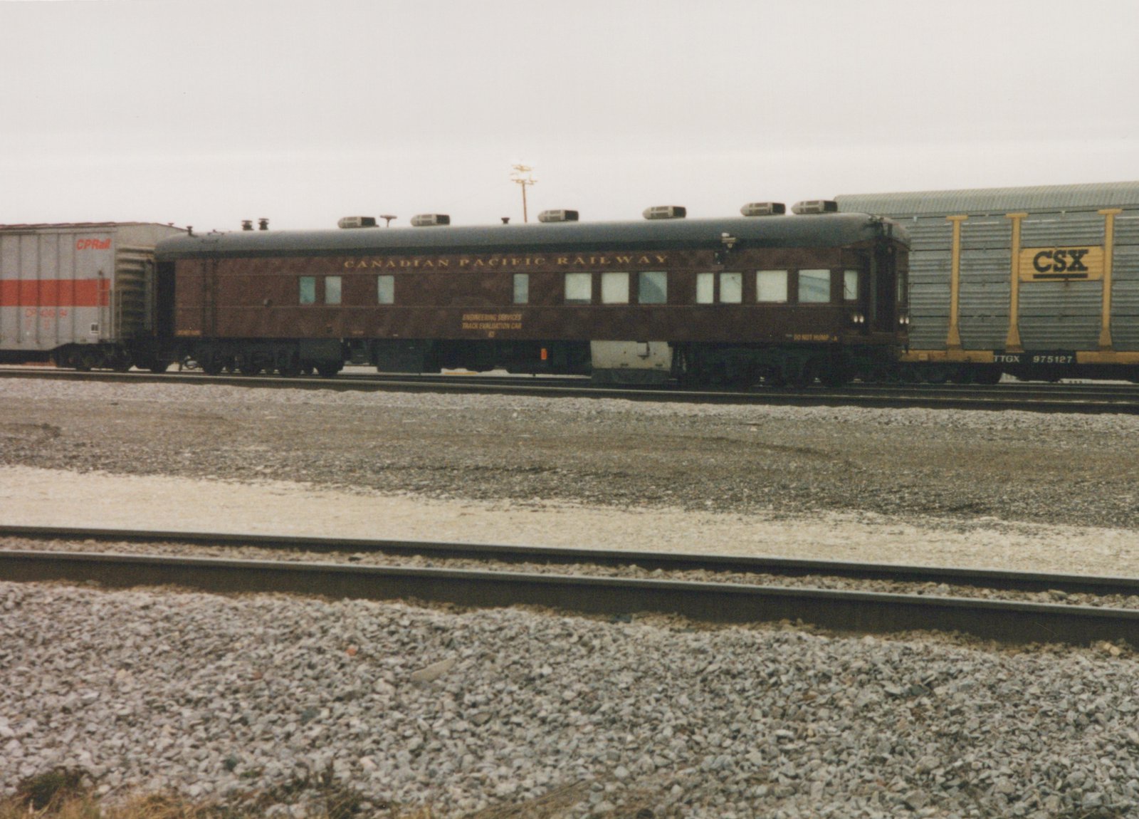 A side view of a brown Canadian Pacific passenger car labeled 'Track Evaluation Car 63' in a gravel rail yard, flanked by a grey CPRail boxcar and a grey CSX freight car under an overcast sky.