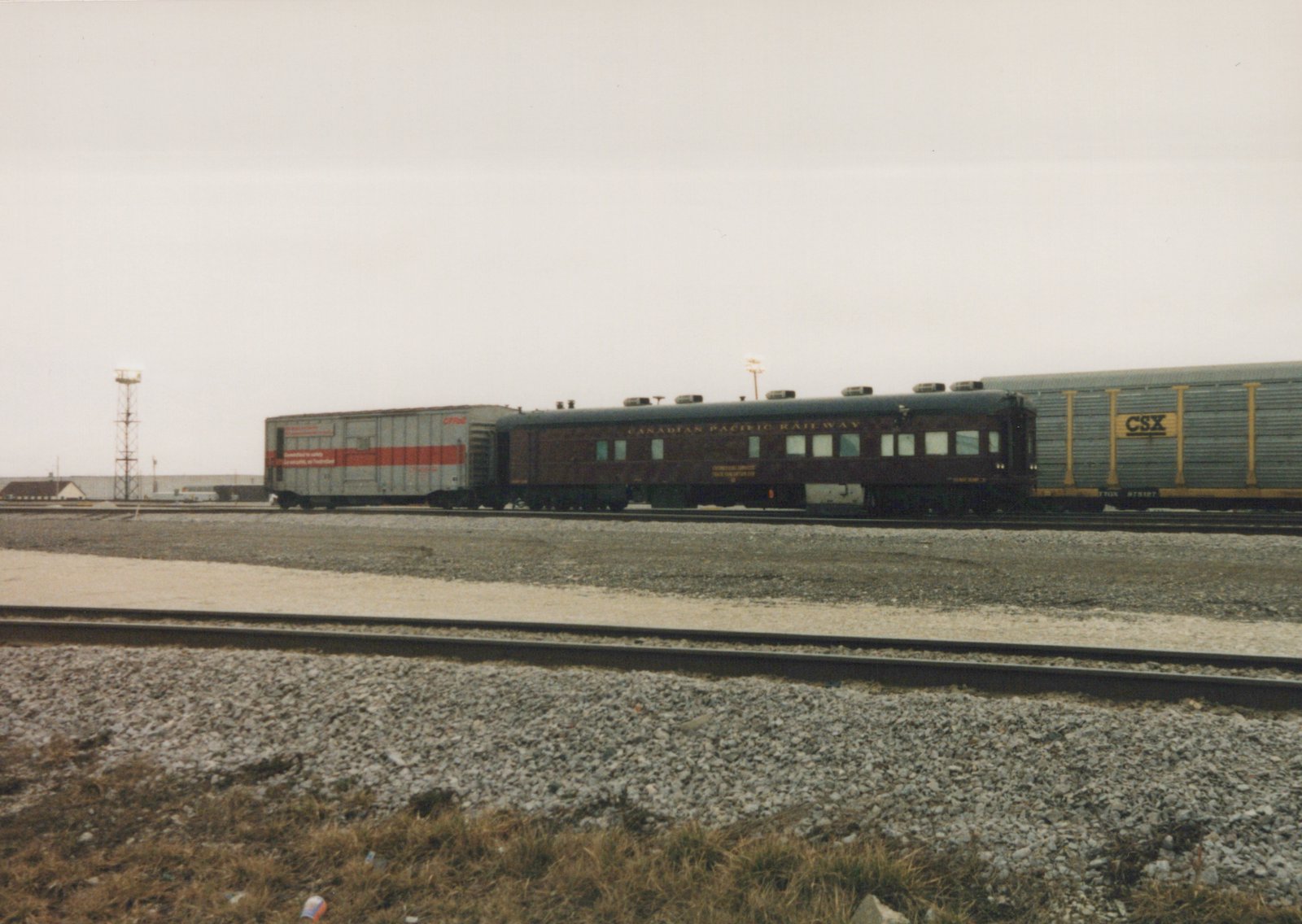 A maroon Canadian Pacific Railway passenger car is parked on a railroad track in a yard next to a silver boxcar and a gray gondola car, all under a gray, overcast sky.