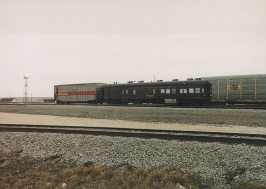 A maroon Canadian Pacific Railway passenger car is parked on a railroad track in a yard next to a silver boxcar and a gray gondola car, all under a gray, overcast sky.