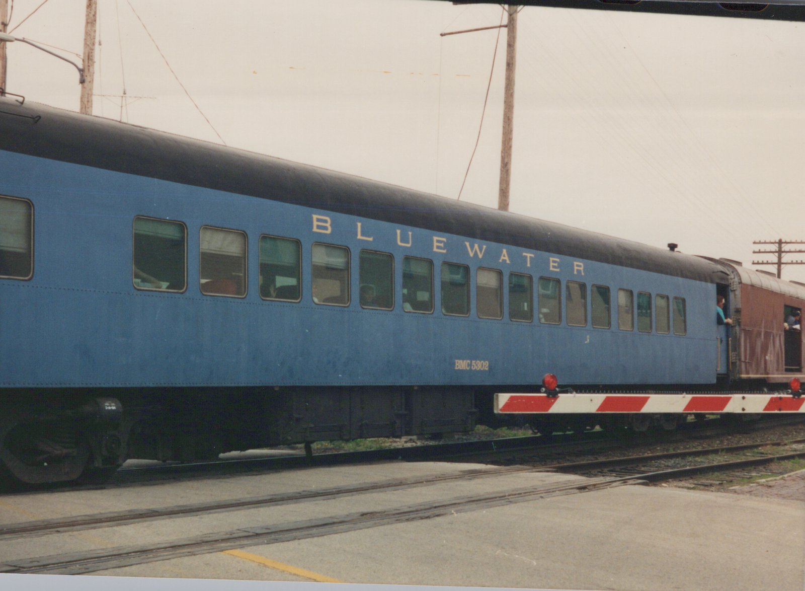 A side view of a bright blue passenger train car with 'BLUEWATER' and 'BMC 5302' written on its side, parked on tracks with a maroon car behind it and people visible in the doorways.