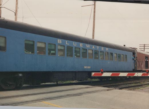 A side view of a bright blue passenger train car with 'BLUEWATER' and 'BMC 5302' written on its side, parked on tracks with a maroon car behind it and people visible in the doorways.