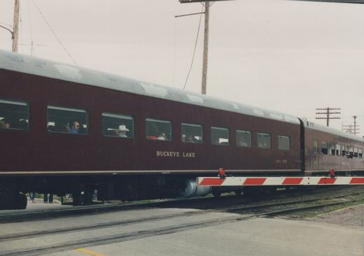 A side view of a two-car passenger train with maroon sides and a silver roof, crossing a street at a grade crossing on an overcast day. The lead car is numbered FWRR 716 and bears the name 'BUCKEYE LAKE'.