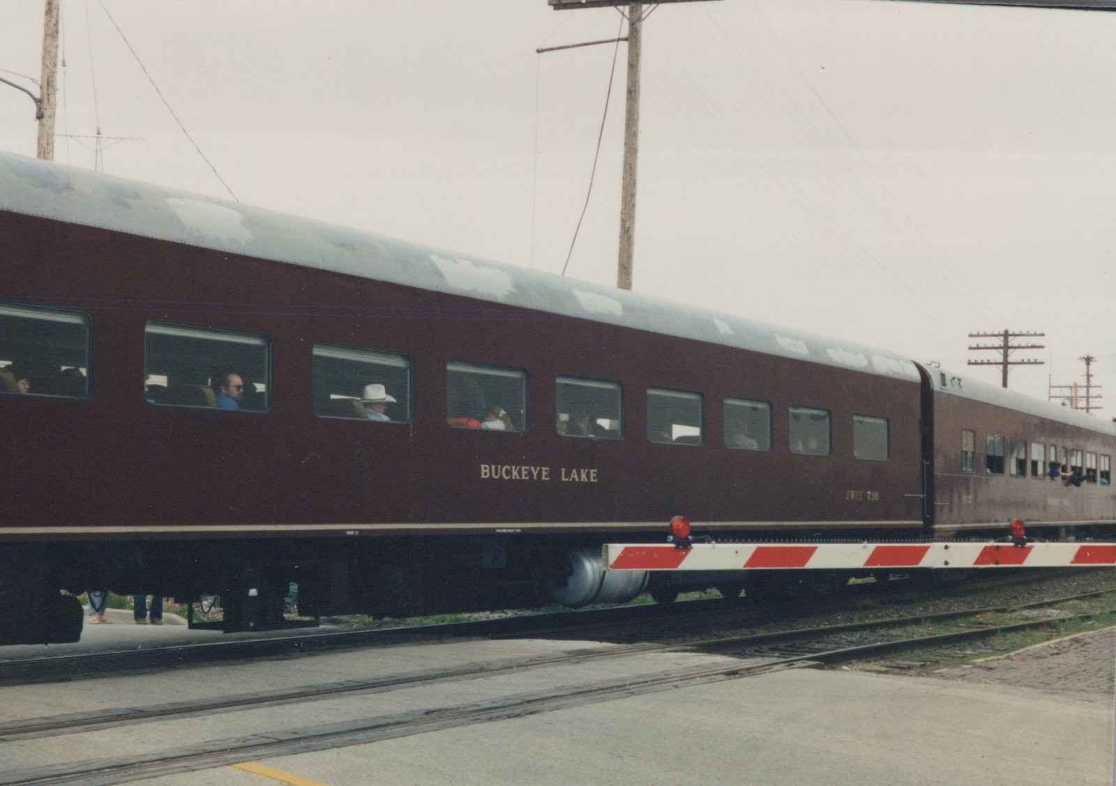 A side view of a two-car passenger train with maroon sides and a silver roof, crossing a street at a grade crossing on an overcast day. The lead car is numbered FWRR 716 and bears the name 'BUCKEYE LAKE'.