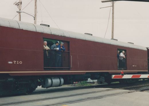 Side view of a maroon passenger train car with the number 710 and the reporting mark FWRH, parked on tracks in a yard with several people leaning out of its end-platform windows.